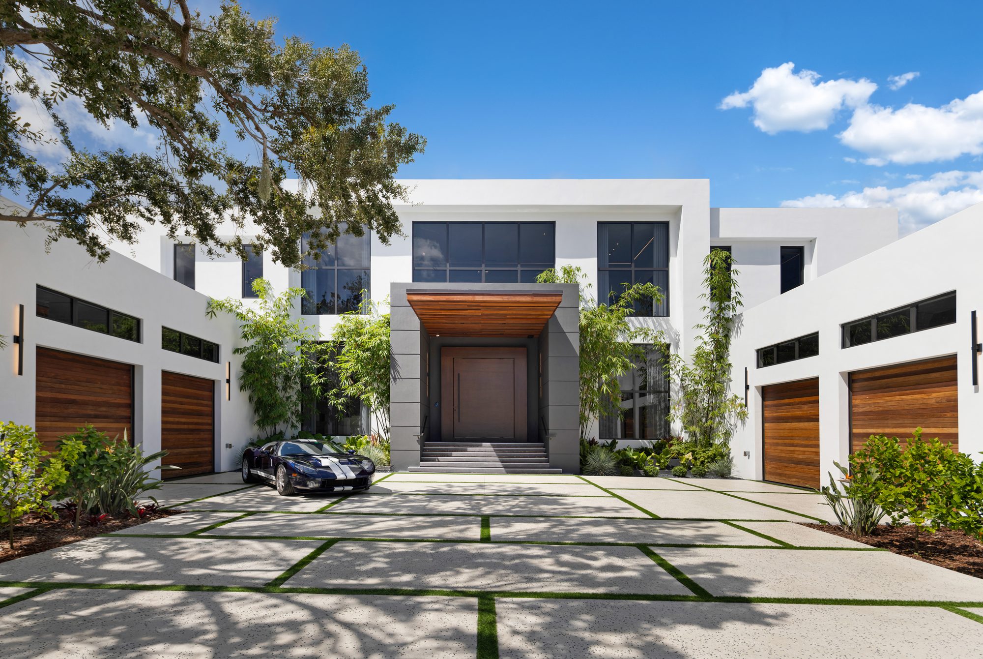 Front elevation of 9900 SW 125th Ave at midday — twin garage wings framing the entry, a heritage-livery Ford GT in the motor court